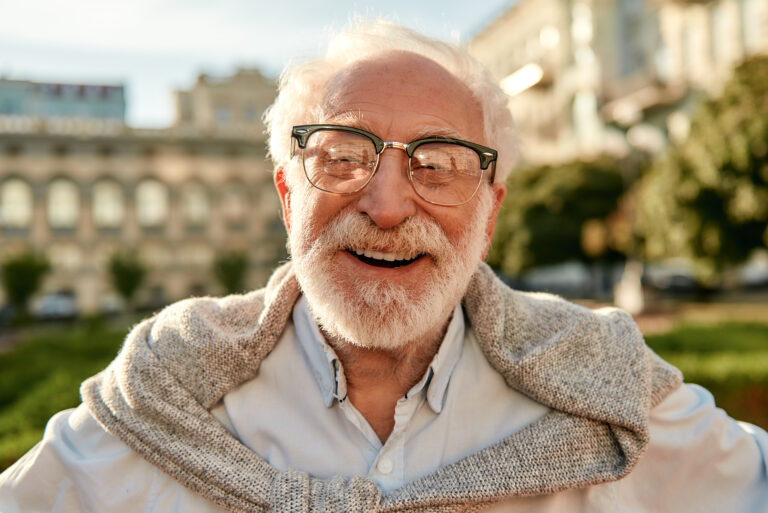 Life Settlement Options- portrait of handsome bearded senior man in glasses looking at camera and smiling while standing outdoors on a sunny day.