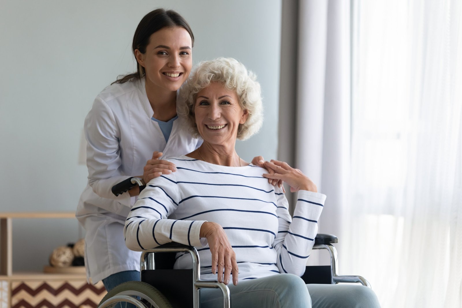 Settlement Services - Senior woman in living room sitting in wheel chair with young nurse smiling looking and looking at the camera. VIATICAL SETTLEMENTS