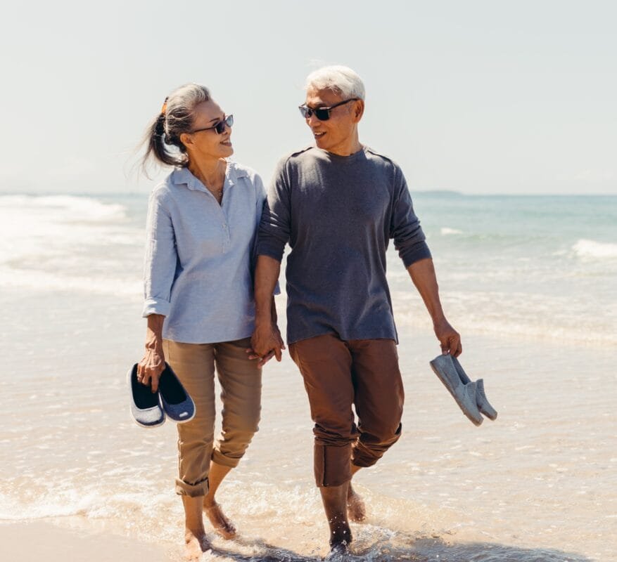 Reverse Life Insurance - Senior couple strolling happily along the beach in the sunshine and bright sky.