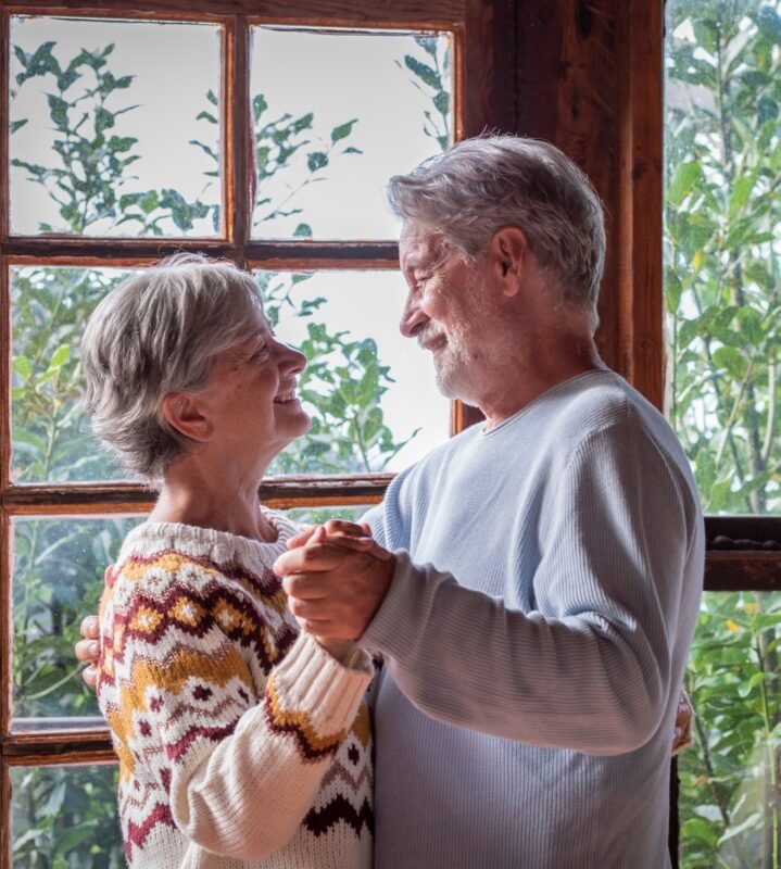 Selling Your Life Insurance Policy - Senior man and woman happy couple dancing at home in living room with outdoors background and windows.