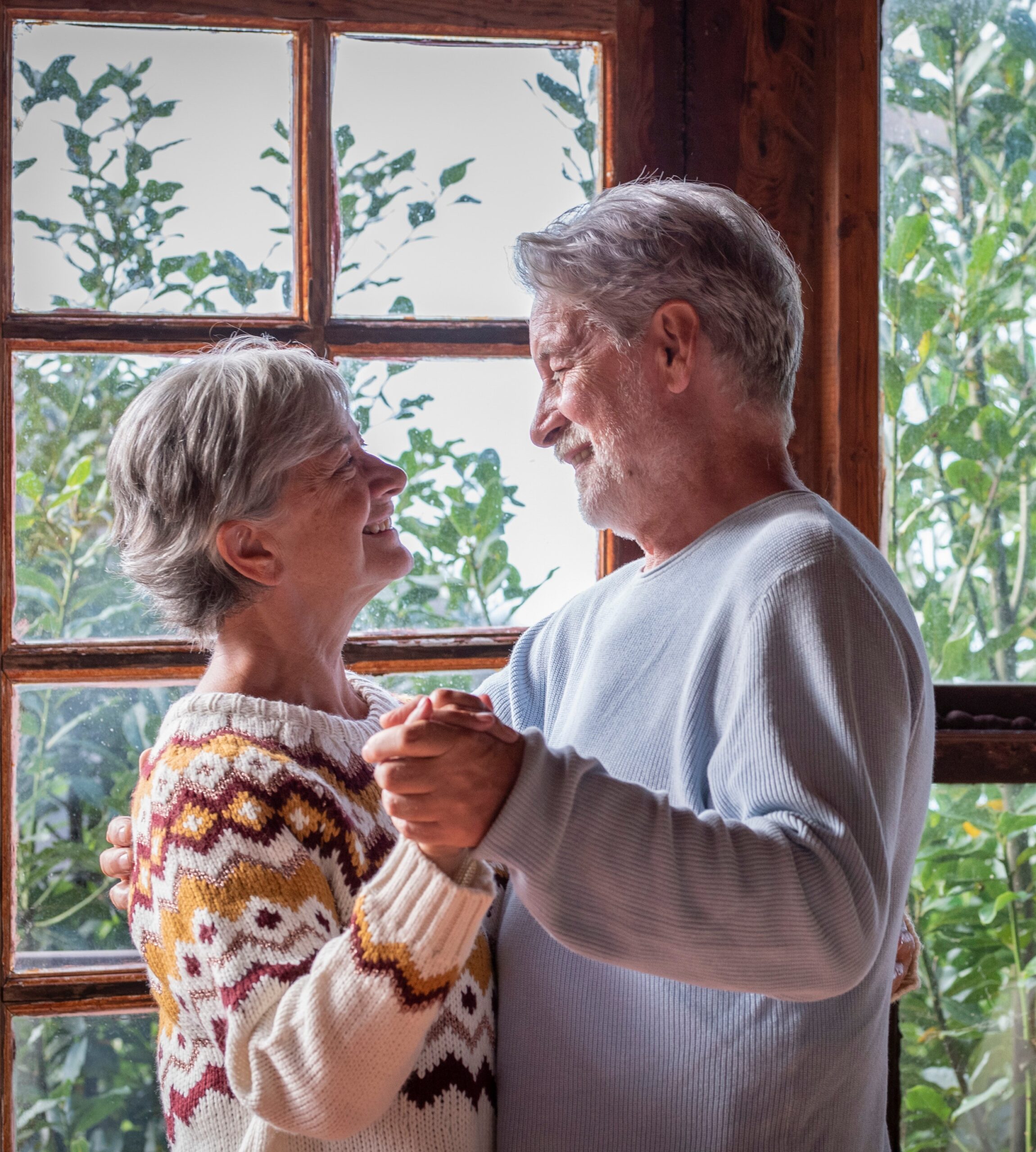Selling Your Life Insurance Policy - Senior man and woman happy couple dancing at home in living room with outdoors background and windows.