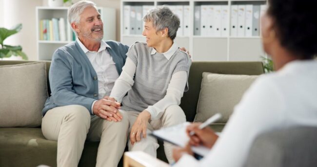 Viatical Settlement vs Life Settlement: Happy senior couple smiling on couch Viatical Settlement vs Life Settlement: Happy senior couple smiling on couch